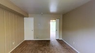This two-floor space presents a clean, functional layout with a neutral design aesthetic throughout. The image showcases a carpeted room finished in warm beige tones, with walls painted in a matching neutral palette that creates a cohesive, understated atmosphere. The flooring consists of brown carpet, adding a layer of warmth and comfort underfoot. White baseboards provide a crisp contrast against the beige walls, contributing to a tidy, well-defined interior finish.

The room features two doorways: one leading to an adjacent space that benefits from natural light filtering through vertical blinds, suggesting an exterior-facing window nearby. A second white door, likely a closet, adds functional storage to the room. The ceiling is smooth and white, maintaining the space's clean and simple character.

The overall ambiance is modest and practical, offering a blank canvas that lends itself well to personalization. The neutral tones and carpeted floors create a quiet, comfortable environment. Spread across two floors, the space provides a multi-level layout suitable for separating living and sleeping areas, making it a versatile and straightforward option for everyday living.