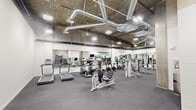 This fitness center offers a comprehensive workout environment spread across a single level, combining industrial architectural character with a full range of modern exercise equipment. The space is defined by its exposed concrete ceilings and visible ductwork, lending an urban, warehouse-inspired aesthetic that contrasts with the clean white walls lining much of the interior.

The main exercise area is generously proportioned, accommodating an array of cardio equipment including treadmills, ellipticals, stationary bikes, and a recumbent cycle. Large wall-to-wall mirrors run along multiple walls, enhancing the sense of depth and allowing users to monitor their form throughout their workouts. Bright overhead lighting ensures the entire space is well illuminated.

A dedicated strength-training section features cable machines, a functional trainer, weight racks, barbells, and adjustable benches, providing options for a full range of resistance exercises. A separate functional training alcove features exposed concrete walls and houses a multi-purpose storage rack stocked with resistance bands, medicine balls, and accessories, along with TRX suspension trainers, offering space for dynamic and bodyweight-focused training. Durable rubberized flooring throughout supports safe, high-impact use across all zones.