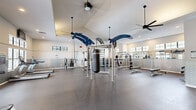 This generously sized fitness center offers a comprehensive workout environment designed to accommodate a wide range of exercise needs. The room features striking vaulted ceilings that create an open, airy atmosphere, complemented by large windows positioned along the upper walls that flood the space with natural light. Multiple ceiling fans provide consistent airflow throughout the room, enhancing comfort during workouts.

The exercise floor is finished with durable rubber flooring, providing excellent traction and impact absorption. At the center of the room stands a prominent multi-functional cable and suspension rig in blue and silver, complete with a heavy punching bag and gymnastic rings — a standout feature suited for functional and combat training. Surrounding it is an extensive selection of strength equipment, including squat racks, weight benches, a barbell station, and a full dumbbell rack.

Along the walls, full-length mirrors amplify the sense of space and allow users to monitor their form. The cardio section is equally well-appointed, featuring treadmills, a StairMaster, and elliptical machines. Tile wainscoting lines the lower walls, adding a polished, clean aesthetic. French doors with glass panes provide access to an adjacent outdoor area, further enhancing the bright and welcoming atmosphere of this facility.