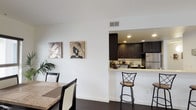 This single-floor residence presents a cohesive contemporary design characterized by crisp white walls, dark hardwood-style flooring, and a neutral color palette maintained throughout. The open-concept living and dining area flows naturally toward a pass-through kitchen, where rich dark espresso cabinetry contrasts sharply with light quartz countertops. The kitchen is well-appointed with stainless steel appliances, including a gas range, built-in microwave, and refrigerator, complemented by ample upper and lower cabinet storage. A breakfast bar with bar seating provides additional casual dining options alongside the dedicated dining space, which features a stone-topped table and receives natural light from a window with blinds.

The unit includes multiple bedrooms, each finished with neutral tones and carpeted or smooth flooring. Bedroom sizes vary, with larger rooms offering space for a queen or king bed alongside additional furnishings, while smaller rooms provide a more intimate layout. Closet access is available in select bedrooms.

The bathrooms maintain the unit's consistent design language, featuring dark vanity cabinetry, light countertops, and wood-tone tile flooring. One bathroom includes a shower-tub combination with sliding glass doors, while another offers a similarly practical layout. Together, the spaces deliver a clean, functional, and modern living environment.