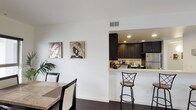 This single-floor residence presents a cohesive contemporary design characterized by crisp white walls, dark hardwood-style flooring, and a neutral color palette maintained throughout. The open-concept living and dining area flows naturally toward a pass-through kitchen, where rich dark espresso cabinetry contrasts sharply with light quartz countertops. The kitchen is well-appointed with stainless steel appliances, including a gas range, built-in microwave, and refrigerator, complemented by ample upper and lower cabinet storage. A breakfast bar with bar seating provides additional casual dining options alongside the dedicated dining space, which features a stone-topped table and receives natural light from a window with blinds.

The unit includes multiple bedrooms, each finished with neutral tones and carpeted or smooth flooring. Bedroom sizes vary, with larger rooms offering space for a queen or king bed alongside additional furnishings, while smaller rooms provide a more intimate layout. Closet access is available in select bedrooms.

The bathrooms maintain the unit's consistent design language, featuring dark vanity cabinetry, light countertops, and wood-tone tile flooring. One bathroom includes a shower-tub combination with sliding glass doors, while another offers a similarly practical layout. Together, the spaces deliver a clean, functional, and modern living environment.