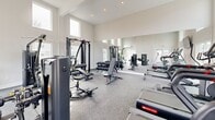 Located at 1000 N Donahue Dr in Auburn, Alabama, this dedicated fitness center offers a comprehensive workout environment within a single, open-plan exercise room. The space is defined by its impressively high ceilings, which create an airy, open atmosphere that makes the room feel more expansive than its footprint suggests. Large windows positioned at multiple heights — including clerestory windows near the ceiling — flood the interior with natural light and provide views of the surrounding outdoor areas, including a pool terrace visible from one side.

The flooring throughout is a durable speckle-finish rubber surface, well-suited for heavy equipment and high-traffic use. The cardio section is well-stocked with treadmills, elliptical trainers, stationary bikes, and additional aerobic machines. A wall-mounted flat-screen TV displaying a fireplace scene adds a welcoming touch to the cardio area.

The strength training section features a robust selection of Hoist brand cable and weight machines, a full dumbbell rack, adjustable benches, and a stability ball. A large floor-to-ceiling mirror along one wall visually expands the space and allows users to monitor their form. Glass double doors lead directly to the outdoor area, enhancing ventilation and accessibility. The overall design is clean, modern, and functional.