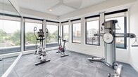 Located at 4555 Minnehaha Ave in the Longfellow neighborhood of Minneapolis, MN, this fitness center offers a well-appointed workout space designed to maximize both functionality and natural light. The room features durable large-format grey rubber tile flooring, ideal for high-impact exercise, and crisp white walls that keep the environment bright and open.

Large, black-framed windows wrap around the space on multiple sides, flooding the interior with abundant natural light and offering elevated views of the surrounding Minneapolis streetscape and neighborhood greenery. Roller shades provide adjustable light control throughout the day. A ceiling fan overhead ensures comfortable air circulation during workouts.

The fitness center is equipped with a range of cardio and strength equipment, including an elliptical trainer, a stationary spin bike, and a multi-function cable resistance machine suitable for a full-body workout. Free weights and a foam roller are also available for supplemental training. A full-length wall mirror amplifies the sense of space and allows users to monitor their form during exercise. A sanitation station is conveniently positioned near the entrance, and posted fitness center rules and workout guides are displayed on the wall. Access is provided through a glass-paneled door, contributing to the open, modern aesthetic of this efficiently designed facility.