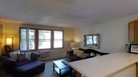 This single-floor space features an open layout that seamlessly connects the living and dining areas. The living room is furnished with plush dark sofas arranged around a sleek black coffee table, creating an inviting seating arrangement. Natural light filters in generously through a set of large windows along one wall, offering views of mature outdoor greenery. A wall-mounted window air conditioning unit and baseboard heating ensure year-round comfort. A decorative framed mirror with an ornate border adorns one wall, adding depth and a classic aesthetic to the room. A warm table lamp provides soft ambient lighting, complementing the natural light. The textured ceiling adds a subtle architectural detail throughout the space. A white dining surface is partially visible, suggesting a dedicated dining area that flows naturally from the living room. The carpeted flooring in a neutral tone ties the space together, contributing to its warm and comfortable atmosphere. Overall, the space offers a functional and welcoming environment with thoughtful layout and classic design elements.