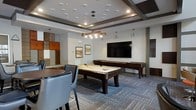 This single-floor amenity space is designed for social gatherings and leisure, combining a modern aesthetic with a welcoming atmosphere. The game room serves as the centerpiece, showcasing a full-size billiards table and an additional shuffleboard table positioned beneath a striking geometric ceiling featuring decorative wood beam accents and a tray ceiling detail. A contemporary chandelier with warm-toned Edison-style bulbs anchors the room, complemented by recessed lighting that ensures even, comfortable illumination throughout. A large flat-screen television mounted on the wall creates a natural focal point, ideal for watching sports or entertainment. The flooring features a patterned area rug in cool blue and grey tones, adding texture and visual interest to the space. Casual seating, including sleek upholstered chairs and a high-top table, is thoughtfully arranged to encourage conversation and relaxation. Framed artwork and wood-paneled wall accents add warmth and a curated, sophisticated touch to the overall design. The space strikes a balance between an upscale lounge and a lively recreation area, making it well-suited for both casual hangouts and organized social events.