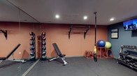 This dedicated fitness space is designed to support a well-rounded workout routine within a compact, efficiently organized layout. The room features warm terracotta-toned walls paired with dark, rubberized flooring, creating a focused and energizing training environment. Recessed ceiling lighting provides consistent illumination throughout the space.

A full-length mirror runs along one wall, enhancing the sense of depth while allowing users to monitor their form during exercise. A wall-mounted pull-up bar offers a sturdy option for bodyweight training, while a TRX suspension system anchored to the ceiling expands functional training possibilities. An adjustable workout bench sits centrally, suitable for a variety of strength exercises.

Along the mirrored wall, a vertical medicine ball rack holds an assortment of weighted balls for dynamic and core training. Additional equipment includes a stability ball, plyo boxes in a bold red finish, and supplementary training accessories. The overall layout is thoughtfully arranged to maximize usable floor space, making this gym practical for solo workouts across a range of fitness disciplines.