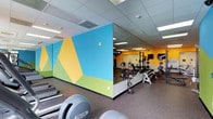 This fitness center is designed to support a comprehensive workout experience across a single, open-plan floor. The space is divided into two distinct zones seamlessly connected by a full-length mirror wall that amplifies the sense of space and enhances visibility throughout. The strength training area is outfitted with Matrix power racks, a full dumbbell rack ranging across multiple weight increments, adjustable benches, and plate-loaded equipment, providing ample options for both beginner and advanced lifters. A dedicated cardio zone, visible along one side of the space, features a row of Precor treadmills and additional cardio machines. The flooring throughout consists of durable, speckled rubber suitable for heavy use and easy maintenance. Overhead, a drop ceiling with recessed fluorescent panel lighting ensures consistent, bright illumination across the entire floor. The walls are finished in bold, color-blocked geometric murals — incorporating shades of yellow, sky blue, and green — creating an energetic and visually dynamic atmosphere. The overall design is clean, functional, and purpose-built to encourage active use.