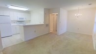 This single-floor space features an open-concept layout that seamlessly connects the kitchen and living area, creating a cohesive and functional floor plan. The kitchen is outfitted with white cabinetry, a built-in microwave, and a peninsula-style counter that doubles as a breakfast bar, separating it from the main living space while maintaining an open sightline. A skylight above the kitchen brings in natural light, brightening the workspace. The walls throughout are painted in a clean, neutral white, contributing to a bright and airy atmosphere. Soft, light-toned carpeting covers the living area, adding warmth underfoot. A doorway visible from the main area leads to additional rooms, with what appears to be a bi-fold closet door hinting at generous storage options. A wall-mounted ventilation unit is positioned near the transition area between spaces. Recessed wall outlets and light switches are placed throughout, and clean white baseboards frame the perimeter of each room. The overall design is simple, functional, and well-maintained, offering a versatile and move-in-ready layout.