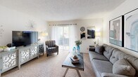 This thoughtfully laid-out single-floor residence offers a complete set of living spaces, including a living room, dining room, kitchen, bedroom, bathroom, and hallway. The carpeted living room is bright and welcoming, featuring a sliding glass door that opens to a private balcony, allowing natural light to fill the space. A cozy seating arrangement and wall art give the room a relaxed, contemporary feel. Adjacent to the living area, the dining room accommodates a round table and chairs beneath recessed lighting, with large windows dressed in sheer curtains adding an airy atmosphere. The galley-style kitchen features warm wood cabinetry, granite-style countertops, a double stainless steel sink, and hardwood flooring, offering efficient use of space with ample storage. The bedroom is a comfortable retreat with plush carpeting, generous natural light through large windows, and room for a full bedroom set. The bathroom is well-appointed with a double-sink vanity topped with granite-style countertops, a tub-shower combination, and classic tile accents. Throughout the residence, a consistent neutral color palette and textured ceilings create a cohesive and inviting ambiance.