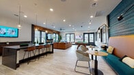 Located at 653 Amherst Rd in Sunderland, Massachusetts, this single-floor community amenity space is thoughtfully designed to blend social, recreational, and collaborative functions in one cohesive environment. The main lounge area serves as the centerpiece, featuring light wood-look flooring, recessed lighting, and a ceiling fan that contribute to a bright and welcoming atmosphere. A striking accent wall clad in warm-toned horizontal wood planks provides a bold visual anchor, complemented by a wall-mounted flat-screen TV and lush indoor greenery throughout. Comfortable seating includes a large sectional leather sofa arranged around a coffee table, alongside casual bistro-style tables and lounge chairs for flexible gathering options. A pool table adds a recreational element to the space, while a teal-painted shiplap accent wall with built-in booth seating introduces a vibrant pop of color. A bar-style counter with pendant lighting and bar stools offers a casual spot for socializing or casual work. Adjacent to the main lounge, private meeting rooms provide a quieter setting, each equipped with a round table, seating for four, a glass whiteboard, and a large interior window with decorative etched glass detailing. These rooms offer views of the surrounding outdoor grounds, keeping the space connected to its natural environment.