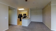 This single-floor residence offers a thoughtfully designed open-concept layout that seamlessly connects the kitchen and living areas. The kitchen is well-appointed with light wood cabinetry, black appliances including a refrigerator, range, and microwave, and a peninsula breakfast bar that provides both additional prep space and a casual dining option. The kitchen flooring features hardwood-style planks, adding warmth and easy maintenance to the culinary space.

Transitioning into the living area, the flooring shifts to plush wall-to-wall carpeting in a neutral tone, creating a comfortable and cozy atmosphere. A decorative pendant light hangs above the dining area, adding a subtle design accent to the open space. Crown molding runs along the ceiling, lending a clean, finished look throughout the unit.

The neutral color palette of soft beiges and whites across the walls and ceilings keeps the space feeling bright and versatile, allowing for a wide range of décor styles. The open flow between the kitchen and living spaces makes the unit feel spacious and functional, ideal for everyday living and casual entertaining. Overall, the space presents a clean, move-in-ready environment with practical finishes and a welcoming ambiance.