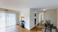 This single-story space is designed with a practical, open layout that promotes easy movement between its main areas. The living area features warm light hardwood flooring that extends throughout, complemented by clean white walls and white baseboards that create a bright, neutral atmosphere. Vertical blind window treatments allow for adjustable natural light from an exterior-facing window, contributing to the room's airy feel.

A partial wall divides the living area from the rest of the unit, providing a sense of definition without fully closing off the space. Beyond this divider, a compact kitchen is visible, outfitted with dark wood cabinetry and essential appliances. Flush-mount ceiling light fixtures provide consistent overhead illumination throughout.

A hallway extends toward the rear of the unit, lined with doors leading to additional rooms. The corridor is well-lit, maintaining the open and accessible feel of the overall layout. White walls and light flooring continue seamlessly from the main living area into the hallway, reinforcing a cohesive and clean aesthetic throughout the entire space.
