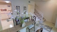 This two-story space combines classic elegance with functional design. The dining area serves as a focal point on the main level, featuring a glass-top table paired with traditional wood chairs with upholstered seats. A striking floral arrangement of white calla lilies adds a refined, decorative touch. The walls are adorned with ornate framed wall art, contributing to the space's sophisticated character. A light-toned area rug defines the dining zone, while tile flooring extends into adjacent areas, including a patterned tile section visible near the staircase. The staircase, finished with white railings and carpeted treads, connects the two levels and adds vertical interest to the open layout. The overall color palette is neutral and light, with white and cream tones creating a clean, airy ambiance. A ceiling vent is visible, indicating a central HVAC system. The open sightlines between the dining area and staircase enhance the sense of spaciousness, making the layout feel connected and cohesive across both floors.