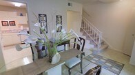 This two-story space combines classic elegance with functional design. The dining area serves as a focal point on the main level, featuring a glass-top table paired with traditional wood chairs with upholstered seats. A striking floral arrangement of white calla lilies adds a refined, decorative touch. The walls are adorned with ornate framed wall art, contributing to the space's sophisticated character. A light-toned area rug defines the dining zone, while tile flooring extends into adjacent areas, including a patterned tile section visible near the staircase. The staircase, finished with white railings and carpeted treads, connects the two levels and adds vertical interest to the open layout. The overall color palette is neutral and light, with white and cream tones creating a clean, airy ambiance. A ceiling vent is visible, indicating a central HVAC system. The open sightlines between the dining area and staircase enhance the sense of spaciousness, making the layout feel connected and cohesive across both floors.