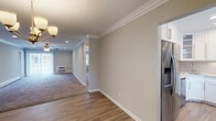 Located at 100 Terrace Road in Bayport, New York, this single-floor residence at Fairfield at Bay Port combines classic architectural details with modern finishes throughout. Crown molding is a consistent design element seen across the living spaces, adding an elegant, refined touch to each room. The open layout flows naturally from the kitchen area through to the living and dining spaces, with a transition from smooth hardwood-style flooring to plush wall-to-wall carpeting that defines the living and bedroom areas. The kitchen is a standout feature, showcasing white shaker-style cabinetry, marble-look quartz countertops, a decorative stone tile backsplash, a stainless steel corner sink with a gold-tone faucet, and a full suite of stainless steel appliances including a gas range, built-in microwave, and French door refrigerator. The living area is generously proportioned, featuring recessed lighting, a ceiling fan, and windows that invite natural light. The bedrooms are calm and comfortable, each finished with soft carpeting and crown molding. The bathroom is bright and clean, featuring a tub and shower combination with sliding glass doors, a marble-top vanity with gold fixtures, and classic basket-weave tile flooring.