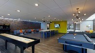 This dynamic single-floor social lounge is thoughtfully designed to serve as a hub for recreation and community engagement. The space features a striking combination of exposed brick accent walls, bold chartreuse-yellow painted surfaces, and warm hardwood-style flooring that together create an energetic yet inviting atmosphere. A drop-panel ceiling with recessed and pendant lighting ensures the entire area is well-lit throughout.

The recreational zone is prominently anchored by a full-size pool table and a shuffleboard table, offering ample opportunity for friendly competition. A wall-mounted flat-screen television is positioned above a long communal high-top table lined with sleek blue metal bar stools, making it ideal for group viewing or collaborative gatherings. Stylish black industrial-style pendant lights hang above this central table, adding a modern design touch.

To one side, a comfortable lounge area features low-profile sofas and vibrant orange accent chairs arranged around small tables, providing a relaxed setting for conversation or casual socializing. Large floor-to-ceiling windows along one wall allow natural light to filter in and offer views of the surrounding exterior, seamlessly connecting the interior to the outdoors. The overall layout is open and spacious, encouraging a variety of uses from active recreation to quiet relaxation.
