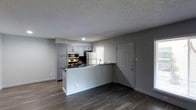 This single-floor space showcases a clean, modern aesthetic built around an open-concept layout that seamlessly integrates the kitchen and living area. The kitchen is defined by a peninsula-style breakfast bar with a white base, providing both workspace and a natural divider between the cooking and living zones. Gray shaker-style cabinetry lines the kitchen walls, offering ample storage, while stainless steel appliances—including a gas range, microwave, and refrigerator—add a contemporary, functional touch. A undermount sink is positioned near a window, allowing natural light to filter into the space. Dark hardwood-style flooring runs throughout, creating a warm contrast against the light gray walls and white trim. The textured ceiling adds subtle visual depth to the interior. A paneled entry door with a deadbolt is conveniently located adjacent to the kitchen, providing direct access to the unit. Overall, the space presents a cohesive, neutral color palette with modern finishes that offer a practical and inviting environment suitable for everyday living.