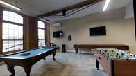 This single-floor game room offers an inviting and energetic atmosphere, blending industrial character with modern amenities. The space is anchored by a full-sized pool table with a classic wooden frame and a blue felt surface, complemented by a foosball table that adds to the recreational variety. Multiple flat-screen televisions are mounted on the walls, ensuring entertainment from every angle in the room. The architectural character of the space is defined by its exposed brick walls and striking rustic wooden ceiling beams, which lend a warm, industrial aesthetic to the environment. An arched window allows natural light to filter into the space, highlighting the textured patchwork-style flooring that runs throughout. A wall-mounted mini-split air conditioning unit ensures year-round comfort. Cue sticks are neatly stored on a standing rack, and additional seating is available for spectators. The overall ambiance strikes a balance between the charm of a historic building and the functionality of a modern recreational facility, making it a well-appointed destination for leisure and social gatherings.