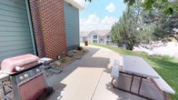 This single-story property includes access to a well-maintained outdoor patio area that serves as a functional communal space. The patio features a smooth concrete surface and is positioned alongside a building constructed with classic red brick and complementary gray-blue siding, giving the exterior a clean, traditional aesthetic. A gas grill is conveniently situated on the patio, making the area well-suited for outdoor cooking and entertaining. A picnic table nearby provides comfortable seating for gatherings. Adjacent to the patio, a small landscaped bed incorporates decorative rock and low-maintenance plantings, adding a touch of greenery without requiring extensive upkeep. A concrete pathway extends from the patio, connecting the space to the broader community grounds. The surrounding landscape includes mature trees that provide natural shade and a sense of privacy. Views from the patio extend outward to neighboring residential buildings and lush green lawns, creating a pleasant community atmosphere. The space benefits from abundant natural light, making it an inviting outdoor retreat during warmer months. Overall, the outdoor area is practical, neatly maintained, and well-integrated into its residential surroundings.