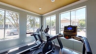 Located at 11655 Briar Forest Dr in Houston, Texas, this single-floor fitness center is a well-appointed exercise facility designed to support a comprehensive workout experience. The space is finished with cool sage-green walls and a warm golden-yellow ceiling accented by crown molding, creating a distinctive and energizing atmosphere. Durable rubber flooring throughout provides a safe, cushioned surface ideal for exercise. Large, multi-pane windows line the walls, flooding the room with abundant natural light and offering views of the surrounding landscape and mature trees outside. The cardio area is well-equipped with multiple treadmills and an elliptical machine, as well as a stationary bike, catering to a variety of aerobic workout preferences. A wall-mounted flat-screen television is positioned for easy viewing during workouts. The space also includes a weight bench and dumbbells for strength training. Glass-paneled interior doors connect the fitness room to an adjacent common area featuring tile flooring, brick accents, and stylish pendant lighting, suggesting a broader community amenity space. The overall design balances functionality with an inviting aesthetic, making it a practical and pleasant environment for daily exercise.