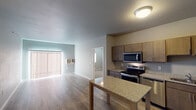 Located at 20th Street South in Brookings, South Dakota, this single-floor unit offers a clean, modern layout designed for comfortable everyday living. The kitchen is a standout feature, equipped with a full suite of stainless steel appliances — including an electric range, microwave, dishwasher, and refrigerator — complemented by warm wood-toned cabinetry, granite-style countertops, and a central island that provides additional prep and dining space. Durable wood-look flooring runs throughout the kitchen and living areas, lending a cohesive and easy-to-maintain aesthetic. The open living area is bright and airy, featuring a sliding glass door with vertical blinds that invites natural light and provides access to the outdoors. The bedroom is spaciously proportioned with plush carpeting, a window with blinds, and a baseboard heating unit. A walk-in closet fitted with wire shelving and hanging rods offers generous storage. The unit includes two bathrooms: one with a full tub-shower combination and granite-topped vanity, and another that connects directly to the walk-in closet. Both bathrooms feature wood-look tile flooring and dark-toned cabinetry with framed mirrors, creating a polished, contemporary finish throughout.