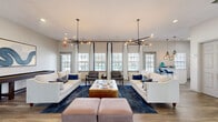 This thoughtfully designed single-floor communal space offers an open and inviting atmosphere across a well-appointed living, dining, and kitchen area. The combined living and dining room serves as the centerpiece, featuring wide-plank hardwood-style flooring, crisp white walls, and an abundance of natural light streaming through multiple large windows dressed with elegant white-and-navy curtains. Two striking sputnik-style chandeliers with warm Edison bulbs anchor the living area, casting a modern yet cozy ambiance over a pair of cream sofas, a gold coffee table, and a navy area rug. A glazed double-door entry offers views to an outdoor pool area, further enhancing the bright, open feel. The dining nook is furnished with a round marble-top table and plush navy chairs, complemented by curated wall art and a console display. The kitchen is efficiently designed with white shaker cabinetry, a subway tile backsplash, quartz countertops, stainless steel appliances including a microwave and beverage refrigerator, and geometric pendant lighting. A breakfast bar with seating provides additional functionality. Throughout, the consistent use of hardwood-style flooring, neutral tones, and contemporary fixtures creates a cohesive and polished aesthetic.