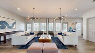 This thoughtfully designed single-floor communal space offers an open and inviting atmosphere across a well-appointed living, dining, and kitchen area. The combined living and dining room serves as the centerpiece, featuring wide-plank hardwood-style flooring, crisp white walls, and an abundance of natural light streaming through multiple large windows dressed with elegant white-and-navy curtains. Two striking sputnik-style chandeliers with warm Edison bulbs anchor the living area, casting a modern yet cozy ambiance over a pair of cream sofas, a gold coffee table, and a navy area rug. A glazed double-door entry offers views to an outdoor pool area, further enhancing the bright, open feel. The dining nook is furnished with a round marble-top table and plush navy chairs, complemented by curated wall art and a console display. The kitchen is efficiently designed with white shaker cabinetry, a subway tile backsplash, quartz countertops, stainless steel appliances including a microwave and beverage refrigerator, and geometric pendant lighting. A breakfast bar with seating provides additional functionality. Throughout, the consistent use of hardwood-style flooring, neutral tones, and contemporary fixtures creates a cohesive and polished aesthetic.