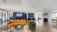 Located at 200 Bicentennial Circle in Sacramento, CA, this single-floor commercial space offers a thoughtfully designed layout that balances open communal areas with private rooms. The expansive main living area immediately impresses with wide-plank hardwood flooring, recessed lighting, and soaring ceilings that amplify the sense of space. Floor-to-ceiling black-framed glass partition walls divide the interior elegantly, creating distinct zones while maintaining visual connectivity throughout. Large windows flood the space with natural light and provide views of the surrounding greenery. The living area is anchored by stylish contemporary furnishings, including a cognac leather sofa and plush green velvet accent chairs, complemented by a bold navy feature wall with built-in shelving and a large-format display screen. A stone fireplace adds warmth near a dining area with bar-height seating, while a glass-enclosed conference room offers a dedicated private workspace. A hallway leads to a compact private room outfitted with a treatment bed and wall-mounted monitor, suitable for wellness or consultation use. The bathroom completes the layout. Throughout, the design blends industrial and modern aesthetics — dark metal accents, neutral tones, and clean lines — creating a polished, professional, and welcoming environment.