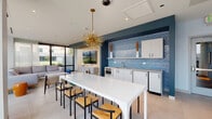 This contemporary single-floor space is thoughtfully designed with an open-concept layout that seamlessly connects the kitchenette, dining, and living areas. The kitchenette is a standout feature, set against a bold teal accent wall adorned with vibrant blue mosaic tile backsplash. White cabinetry, stainless steel appliances, and a sleek countertop complete the functional yet stylish setup. A striking gold Sputnik pendant light anchors the dining area, where a large white table surrounded by mixed seating — including woven rattan and wood-seated chairs — creates an inviting gathering space. Large-format light stone tile flooring runs throughout, lending the space a clean, cohesive feel. The living area is bathed in natural light, courtesy of expansive floor-to-ceiling windows framed in dark metal. Sheer white curtains soften the light while offering sweeping outdoor views, including a cityscape visible from one vantage point and lush greenery from another. A generous sectional sofa in a neutral tone anchors the lounge space, which also features a wall-mounted TV set against warm wood paneling. The overall ambiance is modern, vibrant, and welcoming — a well-curated blend of bold color accents and clean contemporary design.