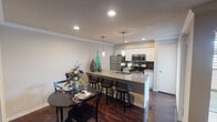 This single-floor residence at 2450 State Hwy 121 in Bedford, Texas, offers a thoughtfully designed layout that balances open communal spaces with private retreats. The kitchen serves as a central highlight, showcasing granite countertop surfaces on both the perimeter and a substantial island with a built-in sink and bar seating. White cabinetry pairs cleanly with stainless steel appliances, including a refrigerator, range, and microwave, while a dark mosaic tile backsplash adds a refined contrast. Pendant lighting above the island enhances the workspace and dining experience.

Rich, dark hardwood flooring flows through the kitchen, dining, and living areas, lending warmth and cohesion throughout. The dining area accommodates a round table with ease, while crown molding adds architectural detail. The living room features a fireplace, windows with natural light, and ample space for seating arrangements.

The bedroom is carpeted and generously proportioned, fitted with a ceiling fan and room for a full furniture suite. The bathroom is finished with granite countertops, white cabinetry, a framed mirror, well-lit vanity, and a tiled tub-shower combination, completing a consistently polished interior aesthetic.