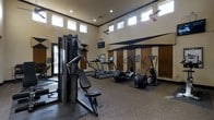 This fitness center is designed to provide a comprehensive workout environment within a single, open-plan space. The room is immediately distinguished by its soaring ceilings, which are lined with a row of clerestory windows along the upper walls, flooding the interior with natural light and creating an airy, open atmosphere. Ceiling fans are mounted overhead to maintain comfortable air circulation throughout the space.

The flooring features a durable, multi-surface layout combining rubberized matting in high-use areas with carpeted sections, offering both safety and comfort underfoot. The walls are finished in a warm neutral tone accented with bold dark geometric striping, giving the space a modern, motivating aesthetic.

The equipment selection is thorough and well-organized. A multi-station cable and weight machine anchors the strength training area, while a dedicated free weights rack provides options for a range of resistance exercises. The cardio zone is equipped with treadmills featuring integrated digital displays, elliptical cross-trainers, and additional low-impact cardio machines, all arranged to allow easy circulation. A wall-mounted flat-screen television is positioned for viewing during workouts. Blinds on the lower windows offer privacy and light control, rounding out this functional and well-appointed fitness facility.