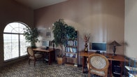 This single-floor space serves as a functional and inviting common area, thoughtfully designed to support productivity and relaxation. The room features a warm, earthy color palette with textured taupe walls that lend a cozy, grounded ambiance throughout. Two dedicated computer workstations are positioned on wooden desks, each accompanied by comfortable upholstered chairs, making the area well-suited for work or browsing. A tall wooden bookshelf stands centrally in the room, filled with a wide selection of books, serving as a focal point for the reading nook atmosphere. Decorative touches are carefully integrated throughout, including tall faux trees that bring a refreshing natural element indoors, along with a decorative vase of floral stems that adds a subtle elegance. The patterned carpeted flooring adds visual texture and warmth to the overall aesthetic. Vaulted wall lines suggest generous ceiling height, contributing to an open and airy feel despite the intimate scale of the room. The space is well-lit, creating a bright and accessible environment. Overall, this area strikes a balance between practicality and comfort, offering residents or visitors a welcoming spot to connect, read, or get work done.