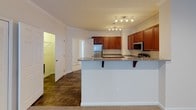 This single-story residence offers a thoughtfully organized layout with multiple rooms visible across its floor plan. The kitchen serves as a functional centerpiece, outfitted with rich medium-toned wood cabinetry, granite-look countertops, stainless steel appliances including a range, microwave, and refrigerator, and light hardwood-style flooring. A peninsula with a breakfast bar overhang connects the kitchen to the adjacent living area, which features crown molding and track lighting overhead.

The bedrooms are carpeted in a neutral beige tone and finished with crisp white trim. Each room benefits from natural light provided by windows fitted with horizontal blinds. Closet space is well-represented throughout, with standard reach-in closets visible in multiple rooms. One bedroom features French doors that open to the exterior, adding both light and outdoor access. Another bedroom includes a vaulted ceiling detail, lending additional visual height to the space.

The bathrooms are finished with wood-look tile flooring and neutral wall tones. One bathroom features a tub-shower combination with a curved curtain rod, while the other includes a tiled tub surround, toilet, and wire shelving for storage. Both bathrooms maintain a clean, functional aesthetic consistent with the rest of the home.