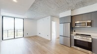 Located at 307 SW 5th St in Fort Lauderdale, FL, this modern one-bedroom, one-bathroom unit showcases a sleek industrial-contemporary design throughout. Exposed concrete ceilings contrast beautifully with crisp white walls and warm light hardwood flooring, creating a refined urban aesthetic that flows through every room.

The well-appointed kitchen features flat-front gray cabinetry reaching to the ceiling, white countertops, a stainless steel sink with a matte black faucet, and a full suite of stainless steel appliances including a refrigerator, range, microwave, and dishwasher. The open layout allows the kitchen to connect seamlessly to the living area.

The bedroom is a comfortable, well-proportioned retreat benefiting from large floor-to-ceiling windows that frame sweeping views of the Fort Lauderdale skyline and surrounding cityscape, filling the space with abundant natural light. The exposed concrete ceiling continues here, reinforcing the unit's industrial character.

The bathroom is finished to a high standard, featuring large-format stone-look wall tiles, a frameless glass-enclosed walk-in shower with mosaic tile flooring, a wall-mounted vanity with an illuminated mirror, and matte black fixtures throughout. A dedicated walk-in closet with wire shelving provides practical storage. The overall ambiance is clean, modern, and sophisticated.