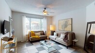 This single-floor residence offers a thoughtfully designed layout that balances functionality with a welcoming ambiance. The living room serves as the heart of the home, featuring wood-style flooring, a ceiling fan with integrated lighting, and generous natural light streaming through large windows dressed with white curtains and blinds. The space flows seamlessly into a dedicated dining area, which is distinguished by a tray ceiling and a pass-through window connecting directly to the kitchen — a practical and stylish architectural detail. The galley-style kitchen is outfitted with white cabinetry, granite countertops, stainless steel appliances including a refrigerator, range, microwave, and dishwasher, and track lighting overhead. A walk-in closet or utility area is visible at the far end of the kitchen. The residence includes two well-proportioned bedrooms, each carpeted and finished with neutral tones, providing a calm and restful atmosphere. Both bathrooms feature white cabinetry with granite countertops, wood-style flooring, and clean, bright finishes; one includes a tub-shower combination with white tile surround. The entry area provides a welcoming transition into the home, complete with a door featuring a sidelight window for added natural light.