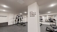 Located at 227 North Walnut in East Orange, NJ, this fitness center offers a dedicated exercise room designed to support a well-rounded workout routine. The space is finished with a clean, modern aesthetic, featuring crisp white walls adorned with motivational phrases such as "Mindset," "Stay Positive, Work Hard, Make It Happen," and "Don't Wish for It, Work for It," creating an energizing atmosphere for users. The flooring throughout is covered in durable rubber gym tile, providing both safety and comfort during exercise. The room is equipped with a variety of strength training machines, including a full power rack with an adjustable bench, a multi-function cable machine, and a red cable/pulley station. Cardio equipment includes multiple treadmills and a stationary bike, complemented by a rack of dumbbells for free-weight training. A large wall-mounted mirror along one side visually expands the space and allows users to monitor their form. The room is climate-controlled with a wall-mounted mini-split unit and is well-lit by recessed ceiling fixtures. Access is provided through sleek black-framed glass doors, and restroom facilities are conveniently indicated nearby. The overall design is functional and motivating, making it an excellent amenity for residents.