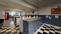 Located at 5820 Murray Dr in Hanahan, South Carolina, this laundromat offers a clean, functional, and well-organized single-floor facility designed to accommodate a high volume of laundry needs. The interior is defined by a striking black-and-white checkered tile floor that runs throughout the entire space, lending a classic, timeless aesthetic to the utilitarian environment.

The facility is generously equipped with rows of commercial-grade front-load and top-load washing machines, as well as stacked dryer units, all featuring modern card or coin-operated payment interfaces with blue payment terminals. Central folding counters with a gray beadboard front provide ample surface area for sorting and folding laundry.

The walls are finished in a two-tone style, with gray beadboard wainscoting below and light gray paint above, creating a clean and cohesive look. Fluorescent lighting ensures the space is brightly illuminated throughout. Windows along the exterior walls allow natural light to filter into the room, contributing to a more open and airy atmosphere.

Seating benches are available near the entrance for patron comfort, and safety equipment including a fire extinguisher is visibly placed. Signage throughout the facility provides instructions and promotes a mobile app for added convenience. The layout is spacious and easy to navigate, making this a practical and well-maintained laundry facility.