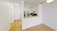 This single-floor residence presents a clean, bright interior with a well-considered layout designed for comfortable everyday living. The living area is a standout space, featuring light wood-style flooring, a classic fireplace framed by crisp white walls, and a large window with vertical blinds that invites ample natural light and provides outdoor views. The open-concept kitchen is efficiently designed with white cabinetry, stainless steel appliances including a full-size refrigerator, a black range, and a peninsula counter with a gray surface that doubles as a breakfast bar. Light wood flooring continues throughout the main living and kitchen areas, creating a cohesive, airy feel. Adjacent to the entry, the layout flows naturally between the kitchen and living spaces. The bedroom offers plush carpeting underfoot, white walls, and a large window with vertical blinds that fills the room with natural light. The bathrooms are finished in white throughout, with one featuring a wall-mounted vanity mirror, globe-style lighting, and an integrated sink countertop, while the second includes a combination tub and shower with sliding glass doors, a toilet, and a mirrored medicine cabinet. A textured ceiling is present throughout the space, adding a subtle architectural detail consistent across all rooms.