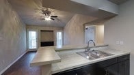 This single-story home showcases an open-concept design that connects the kitchen and living areas into one cohesive space. The kitchen is equipped with a double-basin stainless steel sink, light-toned countertops, and dark cabinetry that provides a striking contrast. A stepped peninsula extends from the kitchen, offering additional workspace and serving as a natural divider between the kitchen and the adjoining living area.

The living room features vaulted ceilings that enhance the sense of openness and volume within the space. A ceiling fan with an integrated light fixture is centrally mounted, providing both air circulation and ambient lighting. The neutral, textured wall finish in a warm taupe tone lends a cohesive and understated aesthetic throughout.

Natural light flows in through multiple windows and a glass-paneled exterior door, brightening the interior and creating an inviting atmosphere. A built-in media niche with dark cabinetry is positioned along the far wall of the living room, adding functional storage and a focal point to the space. Overall, the home offers a practical, well-connected layout with clean finishes and architectural details that add character.