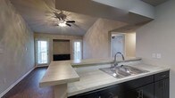 This single-story home showcases an open-concept design that connects the kitchen and living areas into one cohesive space. The kitchen is equipped with a double-basin stainless steel sink, light-toned countertops, and dark cabinetry that provides a striking contrast. A stepped peninsula extends from the kitchen, offering additional workspace and serving as a natural divider between the kitchen and the adjoining living area.

The living room features vaulted ceilings that enhance the sense of openness and volume within the space. A ceiling fan with an integrated light fixture is centrally mounted, providing both air circulation and ambient lighting. The neutral, textured wall finish in a warm taupe tone lends a cohesive and understated aesthetic throughout.

Natural light flows in through multiple windows and a glass-paneled exterior door, brightening the interior and creating an inviting atmosphere. A built-in media niche with dark cabinetry is positioned along the far wall of the living room, adding functional storage and a focal point to the space. Overall, the home offers a practical, well-connected layout with clean finishes and architectural details that add character.