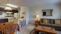 This single-floor residence offers an open-concept layout that seamlessly connects the living area and kitchen, creating a cohesive and functional living space. The living room is carpeted and comfortably furnished, featuring a upholstered sofa accented with decorative pillows, a wooden coffee table, and a side table with a warm lamp that adds a cozy ambiance. A framed mirror with an ornate gold finish adorns the wall, enhancing the sense of space and light. A patterned area rug anchors the seating area, adding visual interest to the neutral-toned room.

The kitchen is positioned at the rear of the space and is partially separated by a half-wall breakfast bar, which is lined with two wooden bar stools. The kitchen is equipped with white appliances, including a refrigerator and range, alongside dark cabinetry that provides a subtle contrast to the light walls. Overhead fluorescent lighting ensures the kitchen is well-lit. The front entry door opens directly into the living space, providing a straightforward and accessible layout. Overall, the unit maintains a simple, practical aesthetic with neutral walls and light-toned carpet throughout.
