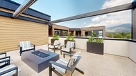This rooftop terrace in Millcreek, Utah offers a well-appointed outdoor retreat with a modern design aesthetic. The space is defined by bold, dark steel pergola beams that frame the open sky above, allowing abundant natural light while providing a sense of architectural structure. The aggregate-finished deck surface extends generously throughout, creating a cohesive and durable foundation for multiple distinct seating zones.

A central lounge area features cushioned armchairs arranged around a cylindrical fire table, ideal for relaxed gatherings. Adjacent to this, a formal dining cluster with woven rattan chairs and a round pedestal table offers a setting suited for outdoor meals or meetings. A third seating arrangement with striped upholstered chairs surrounds a rectangular gas fire pit, complemented by raised planter boxes filled with lush greenery that add color and privacy.

The terrace is enclosed by low parapet walls with large open-frame sections that frame dramatic, unobstructed views of the Wasatch Mountains to the east and the surrounding Millcreek cityscape. Warm wood-panel cladding along the building's facade adds a natural, contemporary contrast to the steel and concrete elements. Sliding glass doors connect the terrace to the interior, ensuring seamless indoor-outdoor flow. This space is well-suited for professional or communal use in an urban setting.