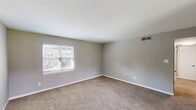 Located at 1220 Carriage House Ln in Gastonia, North Carolina, this well-appointed single-story home offers three bedrooms and three full bathrooms across a generously sized floor plan. The interior is finished with consistent neutral gray wall tones and plush wall-to-wall carpeting throughout the bedroom areas, creating a clean, cohesive aesthetic. The primary bedroom is notably expansive, offering ample space for large furnishings, and features multiple closets with classic panel-style white doors. Additional bedrooms are comfortably sized, each with windows that invite natural light and views of the surrounding neighborhood. A connecting hallway links the bedrooms and bathrooms, providing a logical and functional layout. The bathrooms are outfitted with white vanities, large mirrors, and tub-shower combinations featuring white tile surrounds and wood-look flooring, offering a tidy and practical design. The home includes an entrance that transitions smoothly into the living areas. A covered outdoor area opens to a view of a well-maintained green lawn and neighboring residences, providing a pleasant outdoor retreat. Overall, the home presents a straightforward, functional design with consistent finishes suited for comfortable everyday living.