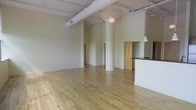 This single-floor loft-style space combines industrial character with modern functionality in an open, airy layout. Soaring ceilings draw the eye upward, where exposed ductwork and conduit run along the ceiling, lending an authentic industrial aesthetic. Polished light hardwood floors extend throughout, reflecting natural and artificial light to create a warm, inviting atmosphere.

Prominent cylindrical structural columns are positioned throughout the main area, adding architectural interest while defining zones within the open floor plan. Pendant lighting fixtures hang at varying heights from the ceiling, providing focused illumination that complements the expansive feel of the space.

A partial-height partition wall on one side hints at a kitchen or bar area, finished with a dark countertop that contrasts elegantly against the light walls and floors. Nearby, a doorway with a warm wood-toned door frame opens to what appears to be a closet or utility area, adding practical storage to the space.

The walls are finished in a clean, neutral tone that enhances the sense of openness and allows the architectural details to take center stage. The overall ambiance is that of a versatile, light-filled loft — well-suited for a variety of uses, from residential living to creative or professional workspaces.