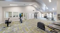 Located at 1700 Hunter Ridge Ln in Peachtree Corners, Georgia, this fitness center offers a generously sized, single-floor layout designed to support a full range of workout activities. The space is defined by its striking vaulted ceilings, which rise dramatically overhead to create an open, airy atmosphere throughout the main exercise area. Flush skylights and recessed fluorescent fixtures ensure consistent, bright illumination across the entire floor.

The flooring features a modern modular carpet tile design in a gray and yellow geometric pattern, adding a contemporary aesthetic while providing comfort underfoot. The main workout area is equipped with multiple treadmills, stationary bikes, an elliptical, a rowing machine, a multi-function cable strength machine, a weight bench, and a dumbbell rack. Wall-mounted mirrors along multiple walls enhance the sense of space and allow users to monitor their form during exercise. A wall-mounted flat-screen television is positioned for easy viewing from the cardio equipment area.

A dedicated TRX suspension training station and additional wall-mounted storage for fitness accessories add functional variety. Natural light filters in through several windows, including a large picture window near the entrance, offering views of the surrounding wooded landscape. A water fountain and glass-panel entry doors complete the facility's practical amenities, making this a comprehensive and well-appointed fitness destination.