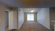 This single-story space presents a clean, functional layout centered around an open living area. The room is finished with wall-to-wall carpeting in a neutral beige tone, complementing the warm-toned walls and white baseboards throughout. A ceiling fan with an integrated light fixture is centrally positioned, providing both air circulation and ambient lighting. A window along the far wall allows natural light to fill the space, creating a bright and welcoming atmosphere. A louvered closet door on the left side of the room offers convenient built-in storage. An HVAC vent is visible near the ceiling, indicating a dedicated climate control system. A secondary doorway on the right side suggests a connection to an adjoining room or entry area, contributing to a practical and straightforward floor plan. The overall design is understated and versatile, with neutral finishes that provide a solid foundation for a variety of interior styles. The space is well-suited for everyday living, offering a comfortable and easily maintained environment.