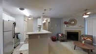 This single-floor space showcases an open-concept design that seamlessly connects the kitchen and living areas, creating a functional and inviting layout. The kitchen features white cabinetry, a peninsula with a light-toned countertop, and three pendant lights hanging overhead, providing both task lighting and visual interest. A closet with bifold doors is visible near the kitchen entry, adding practical storage. The flooring transitions from hardwood-style material in the kitchen area to plush carpet in the living space, defining each zone while maintaining a cohesive aesthetic. The textured ceiling adds dimension throughout. The living area is anchored by a brick-surround fireplace, which serves as a natural focal point. Neutral grey walls create a clean, versatile backdrop throughout the space. Decorative details, including a woven round mirror and ceramic deer head mounted above the fireplace mantle, contribute warmth and character. A hallway visible beyond the kitchen suggests additional rooms, enhancing the sense of a well-organized floor plan. Overall, the space balances practicality with a comfortable, welcoming ambiance.