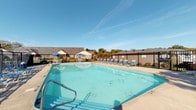 Located at 100 Glenwood Dr in Heath, Ohio, this single-story residential community offers a welcoming and well-appointed living environment designed with comfort and convenience in mind. The property's most prominent shared amenity is a generously sized outdoor swimming pool, enclosed within a black iron perimeter fence that provides both security and an open, airy feel. The pool area is outfitted with a row of blue lounge chairs along the pool deck, inviting residents to relax and enjoy the outdoors. Shaded seating areas with blue umbrellas offer a comfortable retreat from the sun. The surrounding landscape features mature trees that provide a natural backdrop, enhancing the overall ambiance of the outdoor space. The single-floor building design of the community contributes to an accessible, neighborhood-like atmosphere. The exterior architecture reflects a traditional residential style with neutral-toned siding and pitched rooflines visible from the pool area. The combination of thoughtfully arranged outdoor amenities and the calm, suburban setting of Heath, Ohio makes this community a practical and comfortable place to call home.