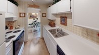 This single-floor residence offers a thoughtfully arranged layout with a warm, traditional aesthetic throughout. The living room provides a comfortable gathering space with neutral carpet, soft wall-mounted lighting, and classic furnishings. Adjacent to it, the dining area features a glass-topped round table beneath an elegant chandelier, with a mirrored wall that amplifies the sense of space and light.

The kitchen is efficiently designed with white cabinetry, a stainless steel sink, light countertops, and plaid wallpaper that adds a cottage-inspired charm. It connects directly to the dining area for convenient flow. A dedicated laundry area houses a stacked washer and dryer unit with built-in shelving for additional storage.

The primary bedroom is generously sized with carpeted floors, a window with decorative curtains, and access to an en-suite bathroom finished with floral wallpaper and a walk-in shower. A second bedroom offers a serene retreat with a walk-in closet and neutral tones. The additional bathroom features deep green accent walls, a framed botanical print, and a tub-shower combination. The office provides a quiet, carpeted workspace with natural light from a large window.