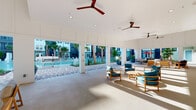 This single-level covered outdoor lounge serves as a central amenity hub, seamlessly blending indoor comfort with open-air living. The space features a clean, modern design with crisp white walls clad in horizontal shiplap paneling and smooth white ceilings fitted with recessed lighting and multiple ceiling fans to ensure comfortable airflow throughout. Large structural columns frame wide, open passages that connect the covered lounge directly to the surrounding pool deck, creating an unobstructed visual and physical flow between the shaded interior and the resort-style outdoor area beyond. The pool deck features a striking herringbone-patterned paver surface, lush tropical palm plantings, and a glass-panel pool surround, all set against the backdrop of a multi-story residential building. Inside the lounge, teak-framed chairs with teal cushions are arranged around low wooden tables, offering a relaxed seating environment ideal for socializing. A separate covered corridor runs along the exterior of the building, lined with blue-trimmed glass doors and windows that allow natural light to filter through while maintaining visual connectivity to the outdoor courtyard. The overall ambiance is bright, airy, and resort-inspired, with a palette of whites, natural wood tones, and coastal blues that reinforces a relaxed, upscale atmosphere.